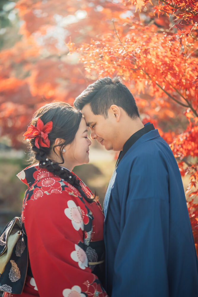 A couple taking photos in front of vibrant red autumn leaves during a romantic Kyoto kimono photoshoot at Maruyama Park