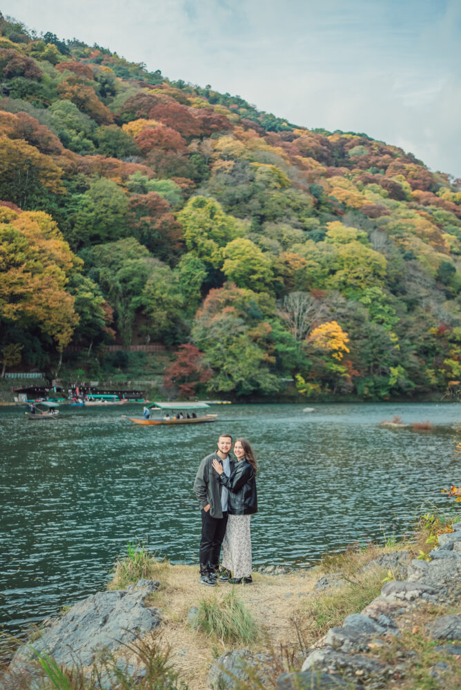 A romantic couple standing by the Hozu-gawa River in Arashiyama Kyoto surrounded by beautiful autumn scenery
