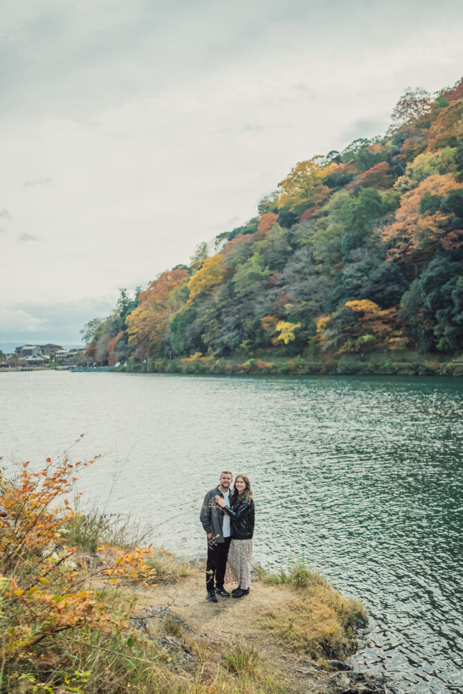 A couple standing along the Hozu-gawa River in Arashiyama Kyoto with colorful autumn leaves and natural scenery in the background