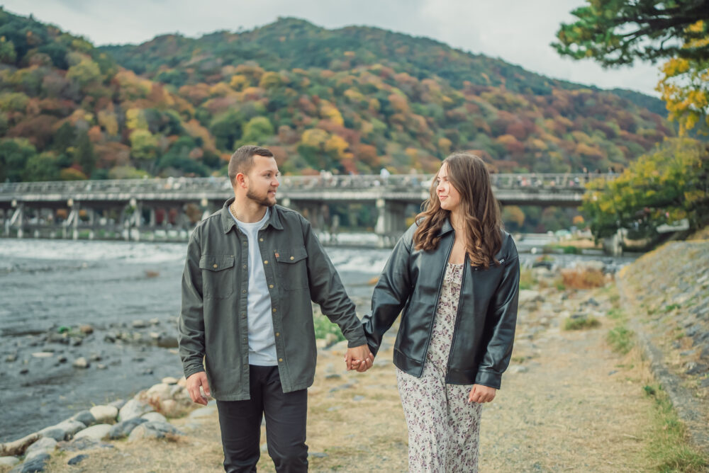 A couple enjoying a romantic moment in Arashiyama Kyoto with Togetsukyo Bridge, mountains, and colorful autumn leaves behind them