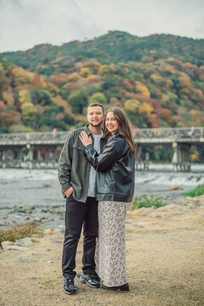 A romantic couple smiling together with Togetsukyo Bridge and beautiful autumn foliage in Arashiyama Kyoto in the background