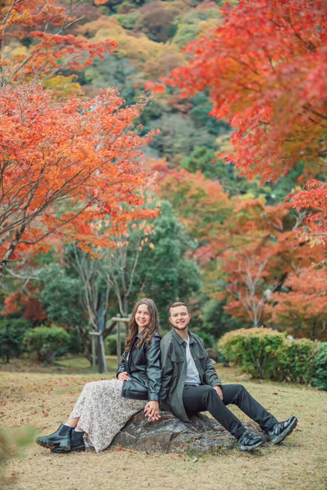 A romantic couple sitting on a stone surrounded by beautiful red autumn leaves in Arashiyama Kyoto during a fall photoshoot
