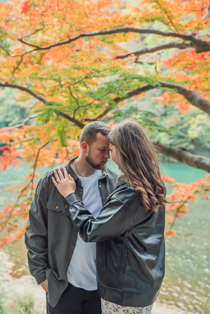 A romantic couple touching foreheads with eyes closed in front of the Hozu-gawa River surrounded by beautiful autumn leaves in Arashiyama Kyoto