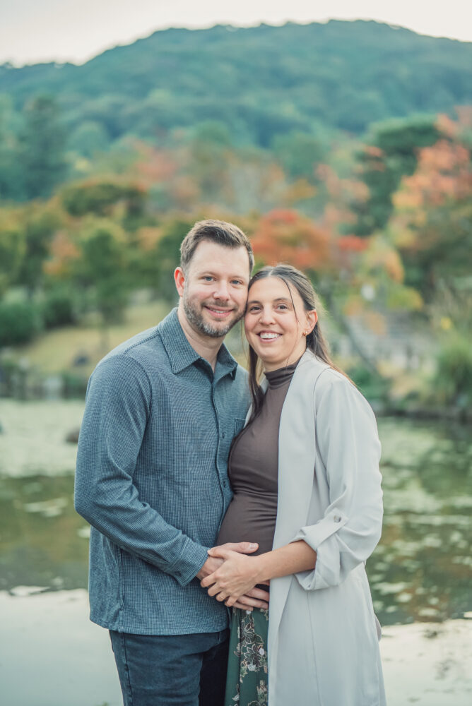 An expectant couple holding hands beside the pond at Maruyama Park with Higashiyama and vibrant autumn foliage behind them during a Kyoto maternity photoshoot