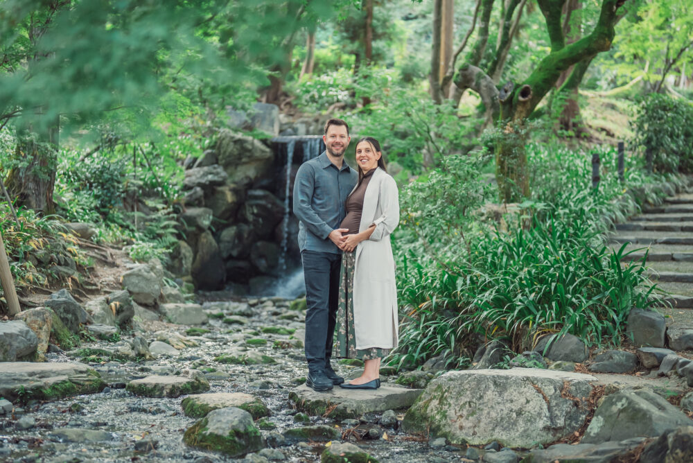 Expecting couple standing on stepping stones in front of a waterfall at Maruyama Park Kyoto during a maternity photoshoot