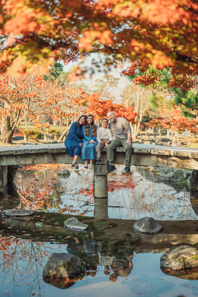 A family sitting on a traditional bridge in Maruyama Park with colorful autumn foliage during a Kyoto family photoshoot
