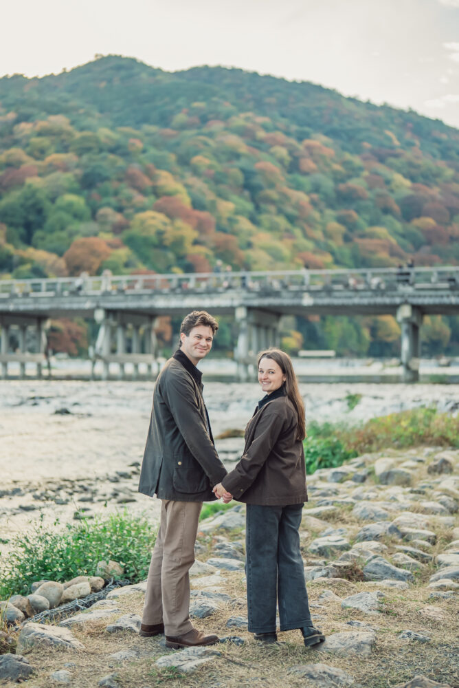 Engaged couple sharing a romantic moment with Togetsukyo Bridge and Arashiyama mountains in the background in Kyoto