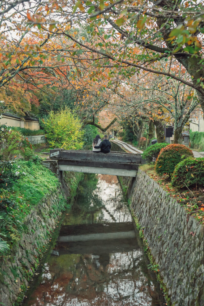 Romantic couple photography at Philosopher's Path Kyoto featuring a small bridge, flowing canal, and colorful autumn foliage