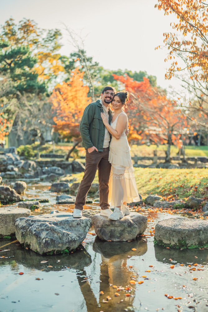 A romantic couple standing on stepping stones over a small stream at Maruyama Park Kyoto surrounded by beautiful autumn leaves