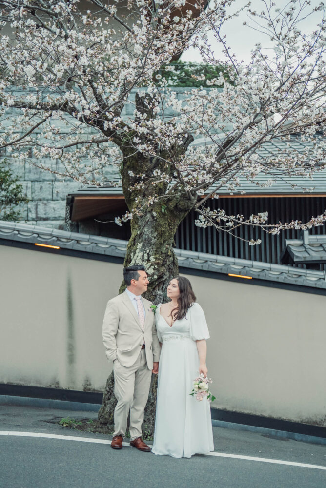 A romantic couple looking at each other beneath cherry blossoms near Yasaka Pagoda in Higashiyama Kyoto during a spring Kyoto elopement and pre wedding photoshoot.