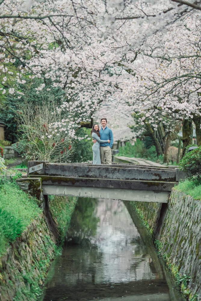 A newly engaged couple standing on a bridge beneath blooming cherry blossoms along Philosopher’s Path in Kyoto after a memorable proposal