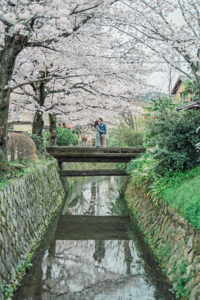 A newly engaged couple standing on a bridge under cherry blossoms along Philosopher’s Path in Kyoto after a surprise proposal