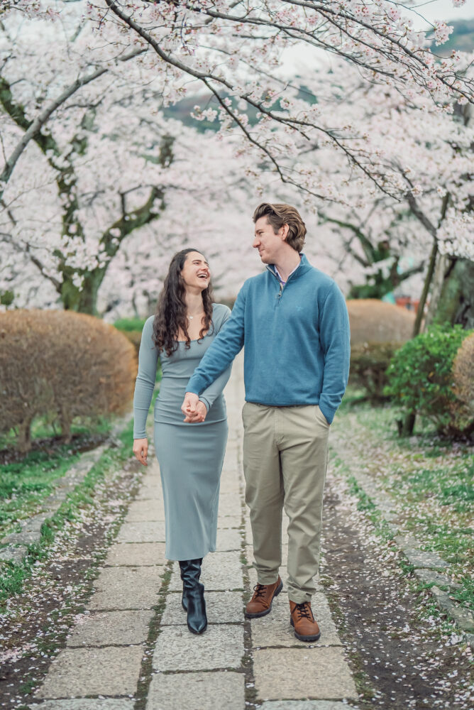 A newly engaged couple walking under blooming cherry blossoms on Philosopher’s Path in Kyoto after a surprise proposal