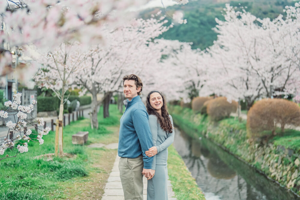 A newly engaged couple standing back to back under blooming cherry blossoms on Philosopher’s Path in Kyoto after a surprise proposal