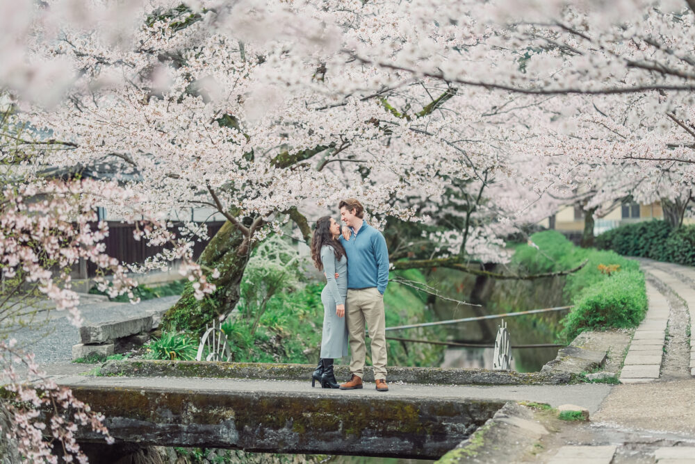A engaged couple sharing a romantic moment after proposal on a small bridge over a canal surrounded by cherry blossoms on Philosopher’s Path in Kyoto