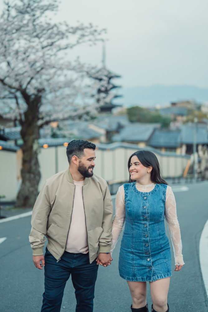 A romantic couple walking hand in hand near Yasaka Pagoda in Higashiyama Kyoto surrounded by cherry blossoms during a spring engagement photoshoot.