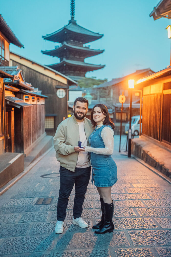 An engaged couple holding a ring box together in front of Yasaka Pagoda in Kyoto during a romantic spring engagement photoshoot.