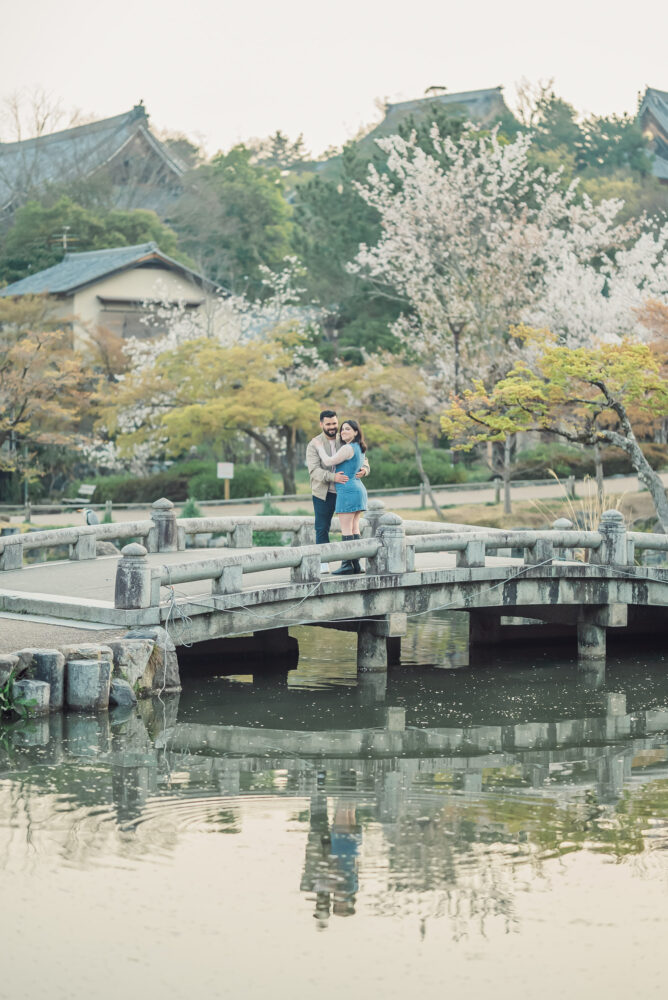 A romantic couple standing on a bridge at Maruyama Park in Kyoto during a sunrise engagement photoshoot surrounded by spring scenery.